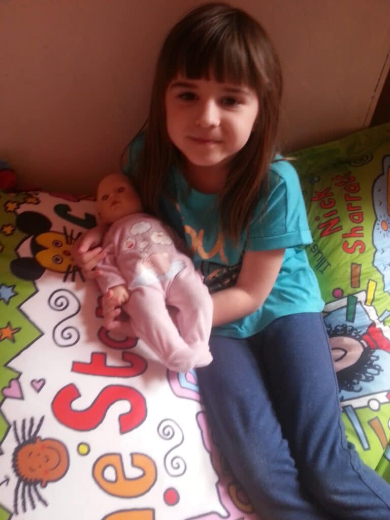 A young girl with straight brown hair, wearing a teal shirt and blue trousers, sits on a colourful bedspread decorated with cartoon characters. She is holding a baby doll dressed in pink pyjamas and smiling softly at the camera.