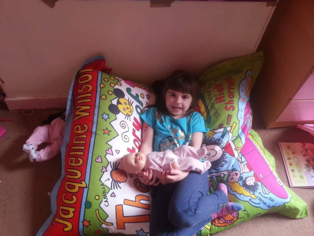 A young girl with brown hair sits on colourful Jacqueline Wilson-themed cushions, smiling and holding a baby doll. She wears a blue T-shirt and jeans. The room has a carpeted floor, some clothes, a book, and a wooden drawer nearby.