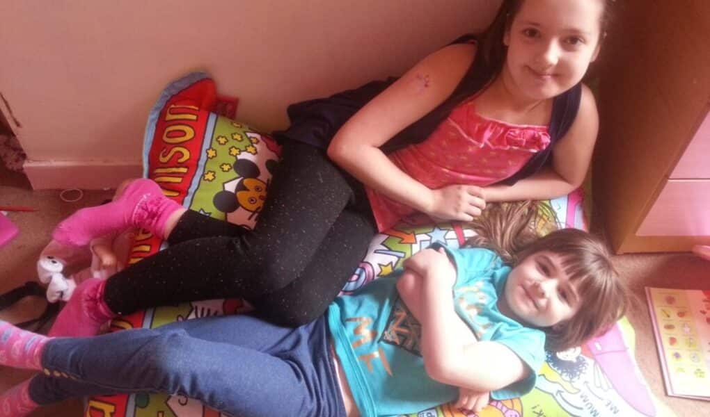 Two young girls lie on a colourful cartoon-themed cushion. The older girl, wearing a pink top and black leggings, props herself on one elbow, while the younger girl in a blue T-shirt and jeans smiles, lying on her back with her arms crossed.