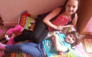Two young girls lie on a colourful cartoon-themed cushion. The older girl, wearing a pink top and black leggings, props herself on one elbow, while the younger girl in a blue T-shirt and jeans smiles, lying on her back with her arms crossed.