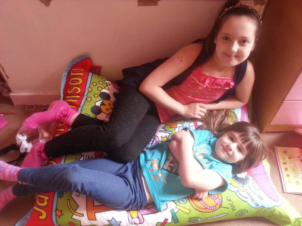Two young girls relax indoors on a colourful cartoon-themed pillow. The older girl, wearing a pink top and black leggings, reclines beside the younger girl, who wears a turquoise shirt and blue trousers. Both are smiling, and the room has toys and books scattered around.
