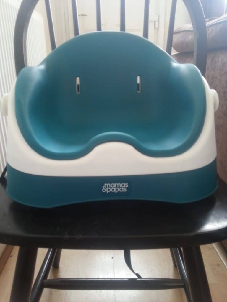 A blue and white plastic booster seat with armrests and the "Mamas & Papas" logo sits on a black wooden chair in a room with wooden flooring, a visible radiator to the left, and a sofa and door in the background.