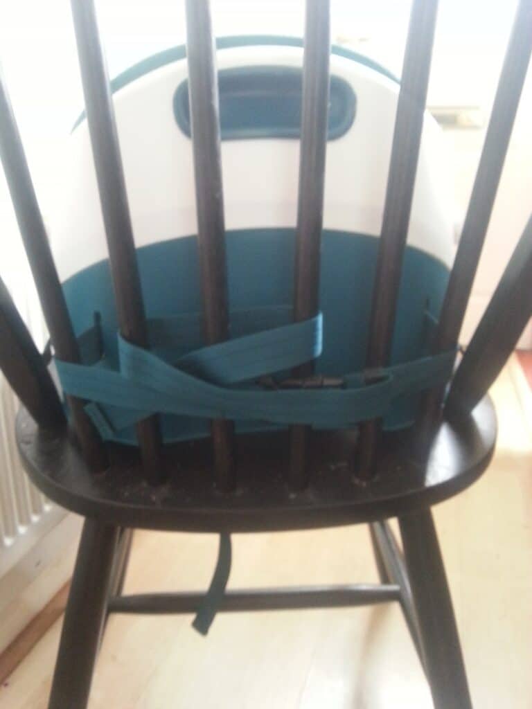 A teal and white booster seat is attached to a dark wooden chair with teal straps threaded through the chair’s back spindles. The image is taken indoors with light coming from a window in the background.