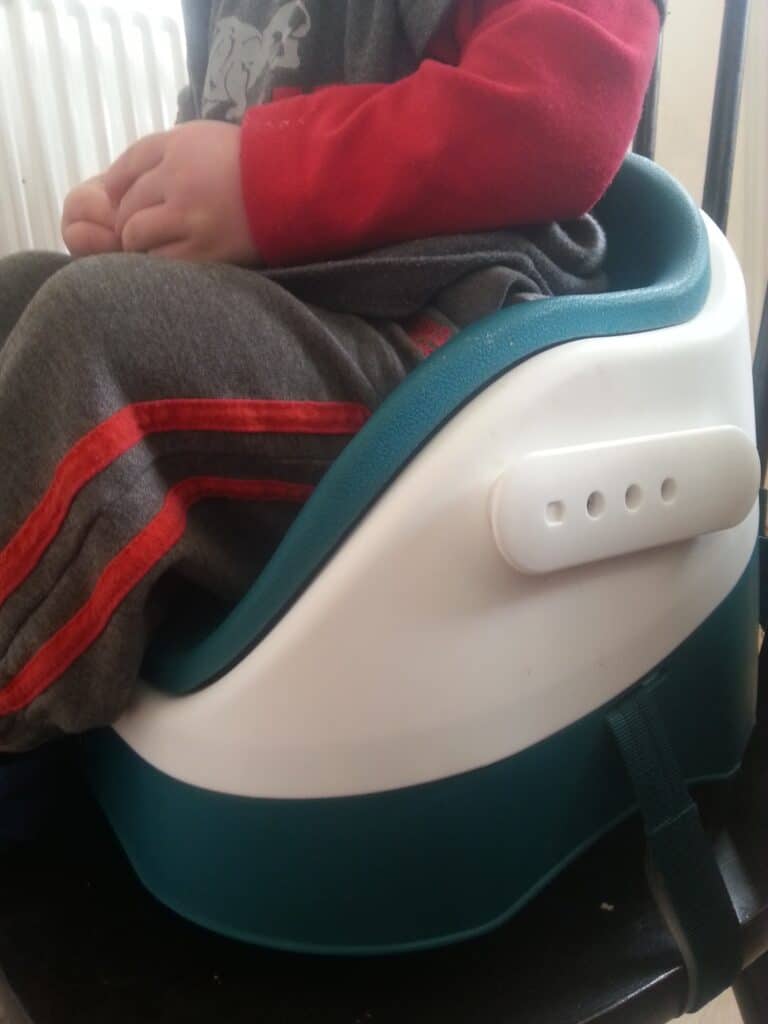 A young child wearing grey trousers with red stripes and a red sleeve sits on a blue and white booster seat attached to a chair. The child’s hands are resting on their lap, and only their lower torso and hands are visible.