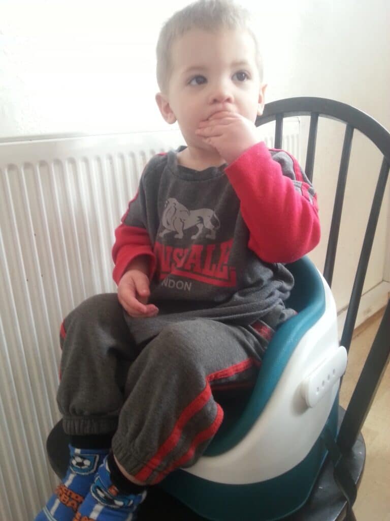 A young child with short blond hair sits on a booster seat placed on a black chair. He wears a grey outfit with red sleeves and stripes, and blue socks with a cartoon character. The child covers his mouth with one hand, looking slightly to the side.