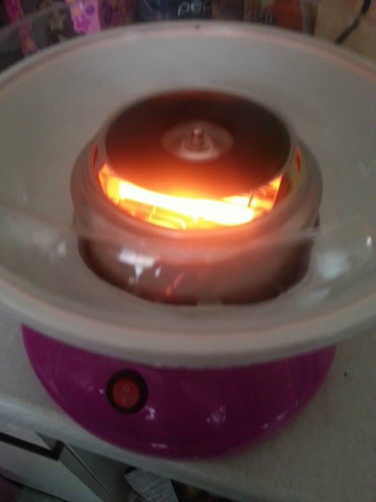 A close-up of a pink candyfloss machine in use, with a glowing orange heating element and spinning head inside, surrounded by a white circular bowl. The power button is visible on the front of the machine.
