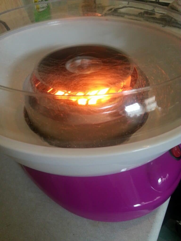 A pink and white candyfloss machine in use, with spun sugar forming around the centre as the machine glows orange from the heating element. The clear plastic lid allows the spinning sugar to be visible. The device rests on a light-coloured worktop.