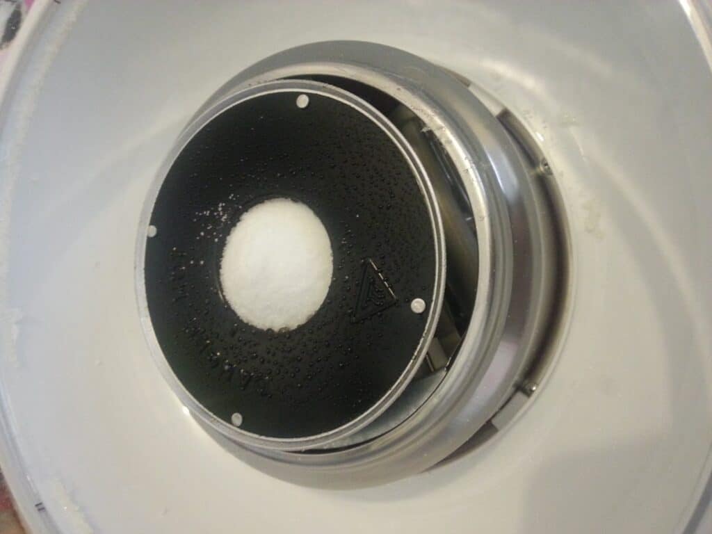 A close-up of a wet, black circular surface inside a metallic appliance, possibly an ice cream maker. There is a mound of white granulated substance, likely salt, in the centre of the circle, surrounded by stainless steel parts.