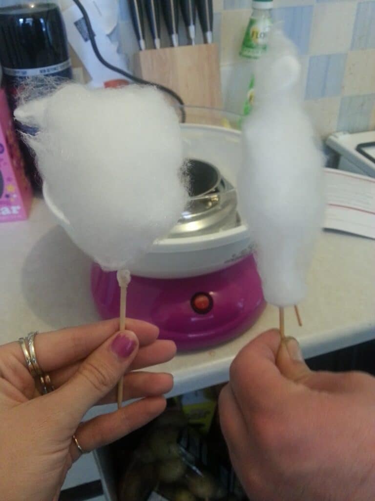 Two hands hold sticks of homemade candyfloss in front of a pink candyfloss machine on a kitchen worktop. One stick has a fluffy, uneven candy, while the other is more wispy and tapered. Kitchen items and knives are visible in the background.