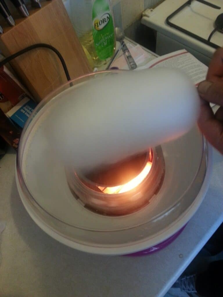 A person spins a stick to collect white candyfloss forming from a machine. The machine's inner chamber glows orange, indicating it's on. In the background, there's a wooden knife block, Flora washing-up liquid, and part of a gas cooker.