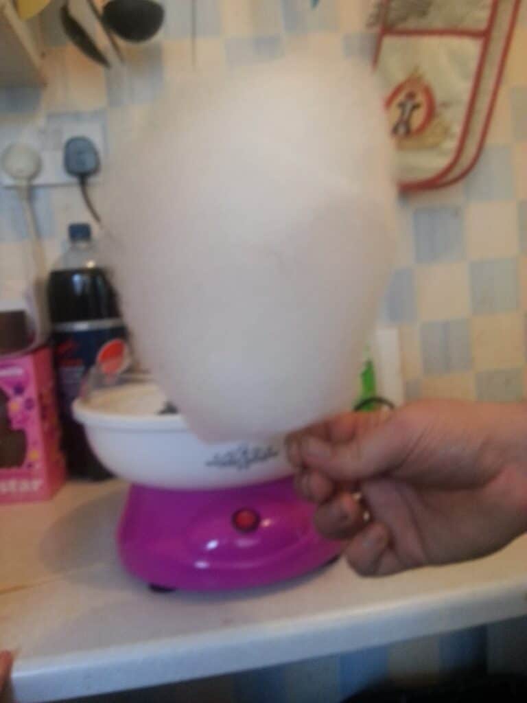 A hand holds a stick of white candyfloss in front of a pink and white candyfloss machine on a kitchen worktop. In the background are a drinks bottle, a pink box, power sockets, and a hanging oven glove on a tiled wall.