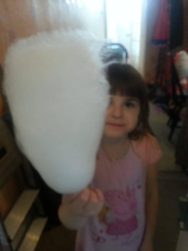 A young girl with brown hair, wearing a pink Peppa Pig shirt, holds a large stick of white candyfloss in front of her. The candyfloss is close to the camera, making it appear oversized and slightly out of focus. The background is indoors and blurry.
