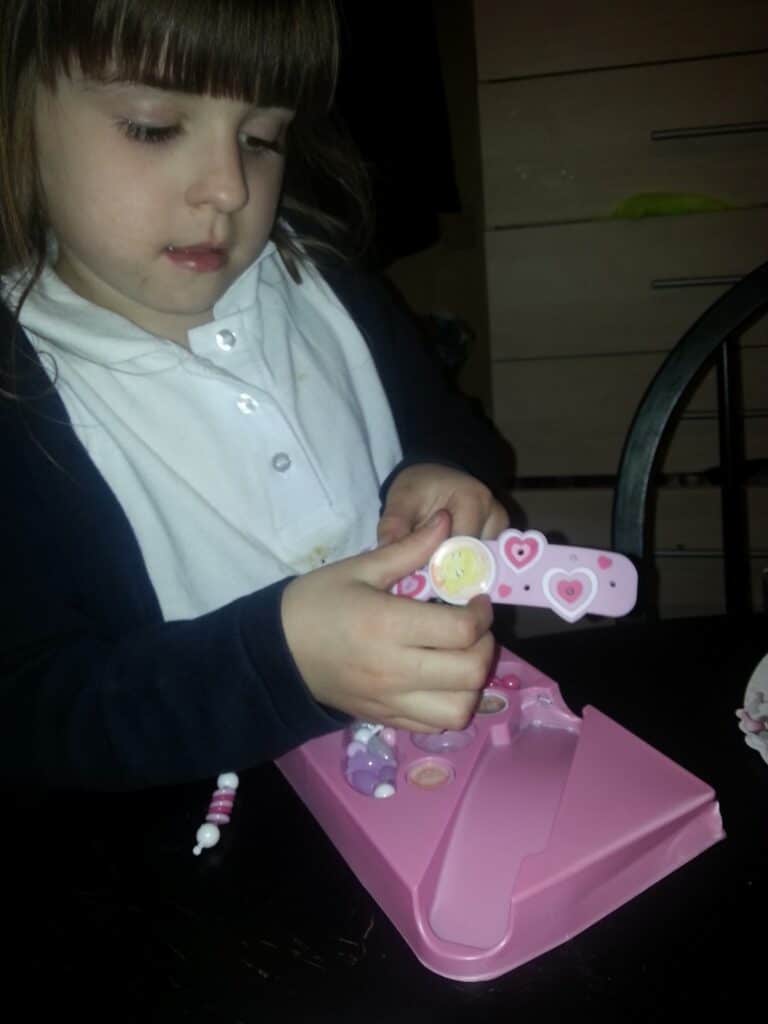 A young girl with light brown hair and fringe, wearing a white shirt and dark jacket, sits at a table using a pink nail file decorated with hearts. She is focused on filing her nails, and a pink case with nail accessories is on the table in front of her.