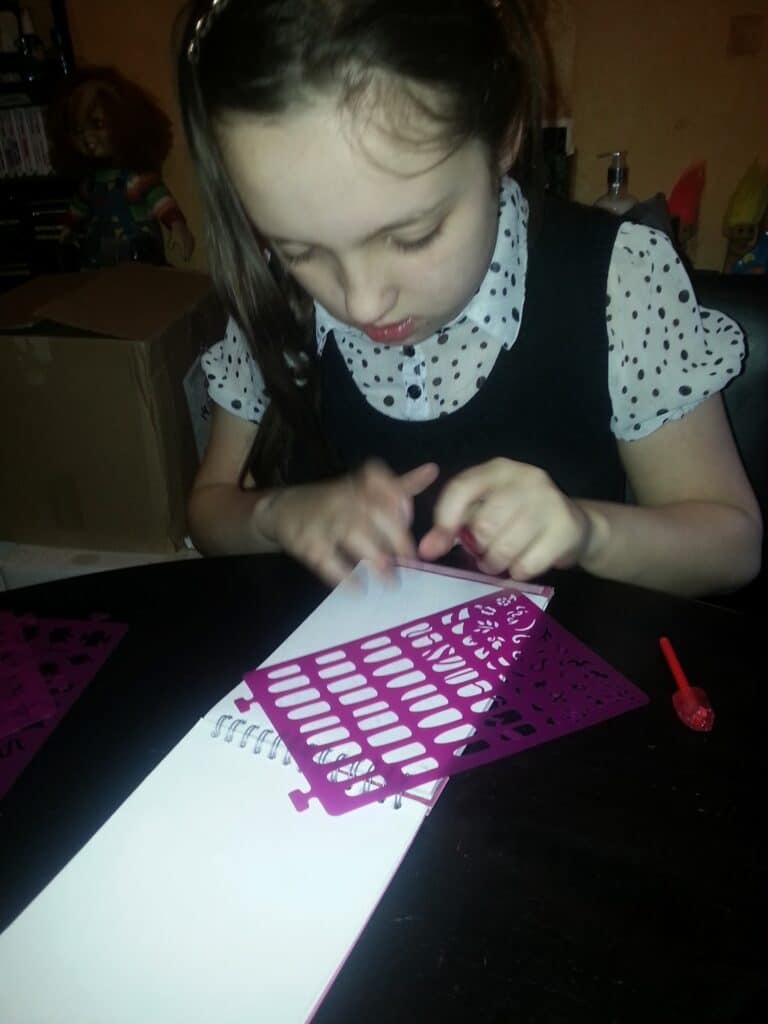 A young girl in a polka-dotted blouse and dark dress focuses intently on tracing stencil shapes onto white paper using a pink stencil ruler. Art supplies and a doll are visible in the background on a dark tabletop.