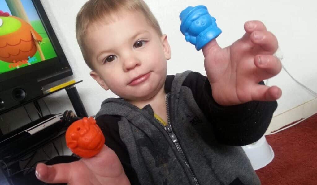 A young child in a grey and black hoodie smiles softly while holding up two colourful plastic finger puppets—one orange and one blue—in front of a TV showing a cartoon on a black TV stand, with a white wall and red carpet in the background.