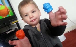 A young child in a grey and black hoodie smiles softly while holding up two colourful plastic finger puppets—one orange and one blue—in front of a TV showing a cartoon on a black TV stand, with a white wall and red carpet in the background.