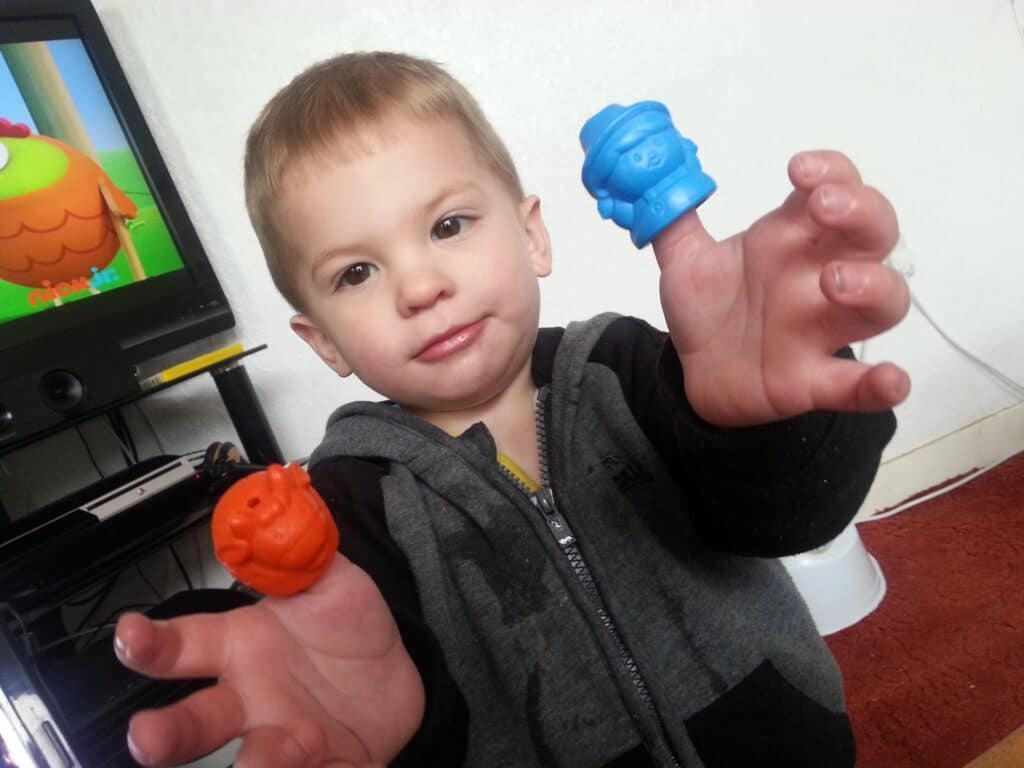 A young child in a grey and black hoodie smiles at the camera, holding up both hands with a blue toy on one finger and an orange toy on the other. A TV and various household items appear in the background.