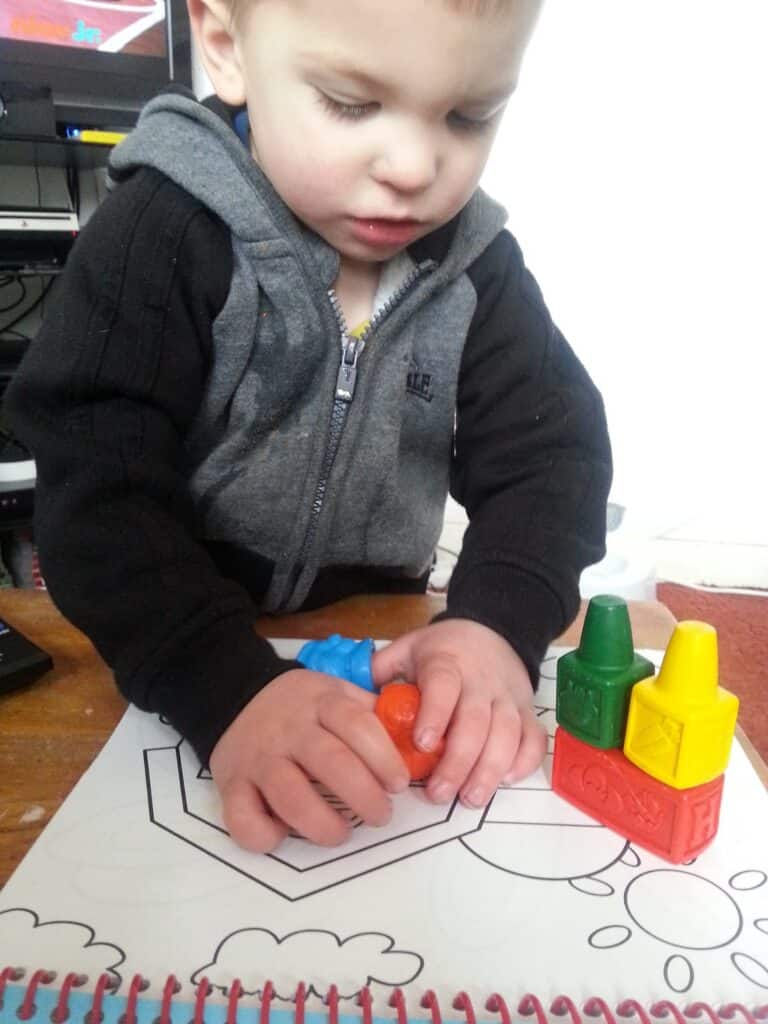 A young child in a grey and black hoodie colours in a colouring book using large orange and blue crayons. Three other crayons—yellow, green, and red—are stacked nearby on the wooden table. The child looks focused on their colouring activity.