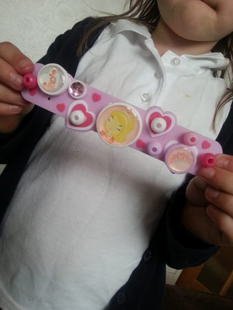 A child in a white polo shirt and navy jumper holds a pink bracelet decorated with heart shapes, pink spots, and round buttons, including one with a cartoon girl's face. The child’s face is partially out of the frame.