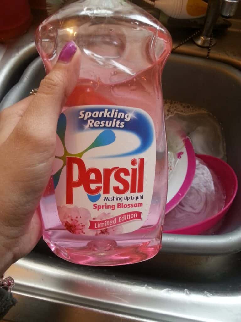 A hand with painted nails holds a clear pink bottle of Persil Washing Up Liquid, labelled “Spring Blossom” and “Limited Edition.” In the background, a sink contains water, dirty dishes, and a pink bowl.