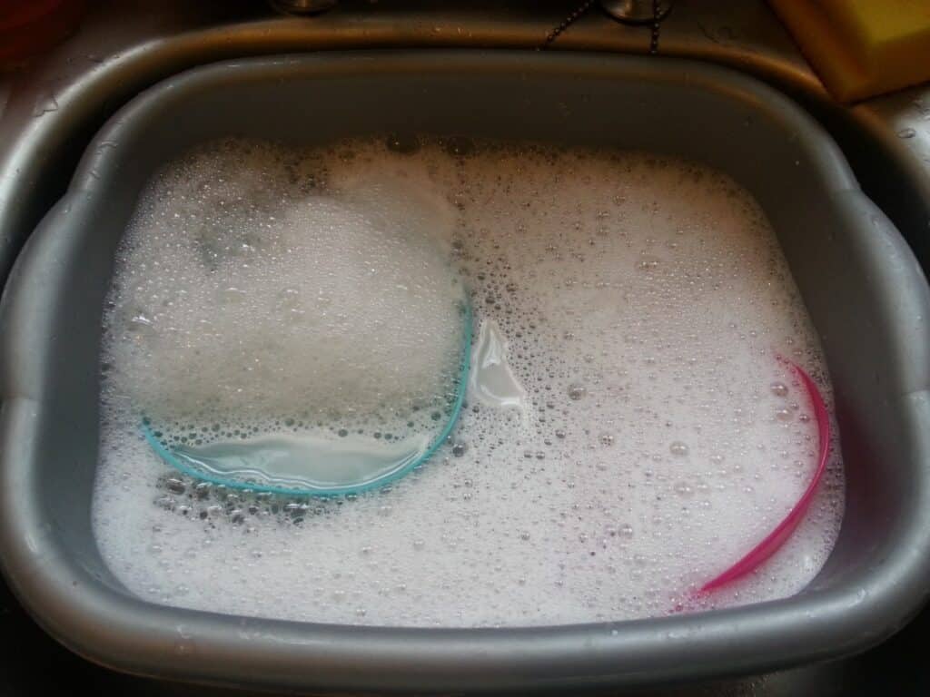 A grey plastic washing-up bowl filled with soapy water and white bubbles; two translucent plastic bowls, one blue and one pink, are submerged and partially visible. The washing-up bowl sits in a metal sink with a yellow sponge in the corner.
