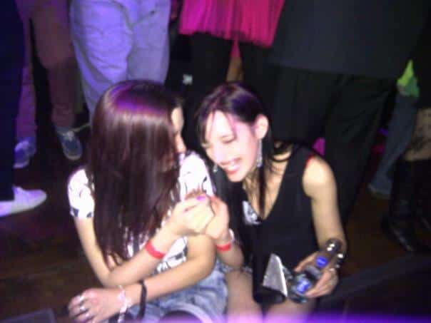 Two young women sit on the floor at a crowded party or club, smiling and laughing together. One holds a drinks bottle, and they appear to be sharing a joyful moment. Colourful lights and people standing around them suggest a lively atmosphere.