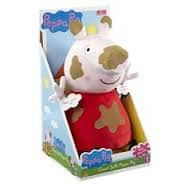 A Peppa Pig soft toy with brown muddy spots wears a red dress and sits upright in an open display box decorated with clouds and grass. The soft character is smiling, with one arm raised, inside the packaging.