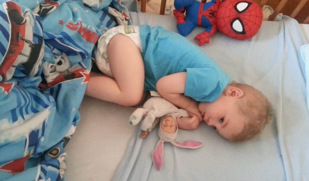 A young child in a blue shirt and nappy lies curled up in a cot, cuddling a soft toy in bunny pyjamas. A plush Spider-Man toy rests nearby. The bedding features cars and aeroplanes on a blue background. The child looks peaceful and comfortable.