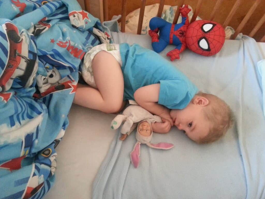 A young child wearing a blue shirt and nappy lies curled up on a bed, cuddling a soft doll in a bunny outfit. The child rests on a light blue sheet under a blanket with aeroplanes. A Spiderman soft toy sits near their head by the wooden cot bars.