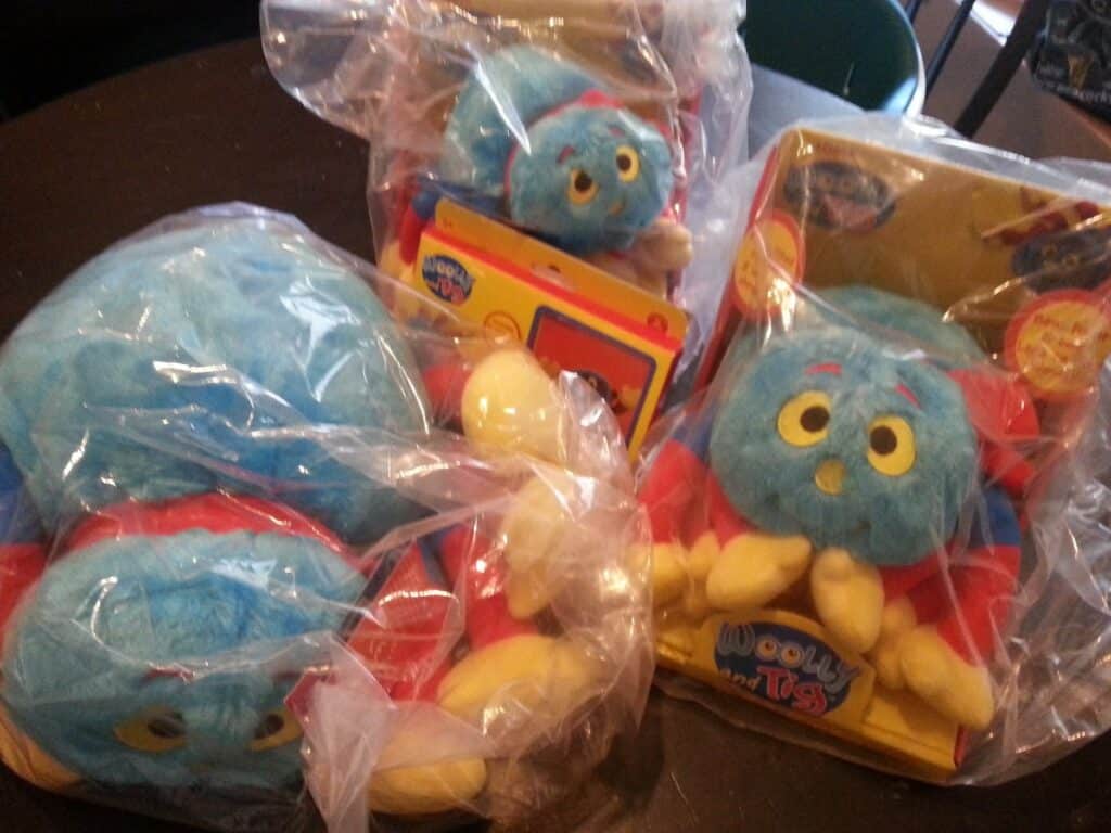 Three blue, red, and yellow plush spider toys are shown; two are in clear plastic bags, and one is in packaging labelled “Wooly and Toy.” The spiders have big, yellow eyes and six yellow legs each, sitting on a dark surface.