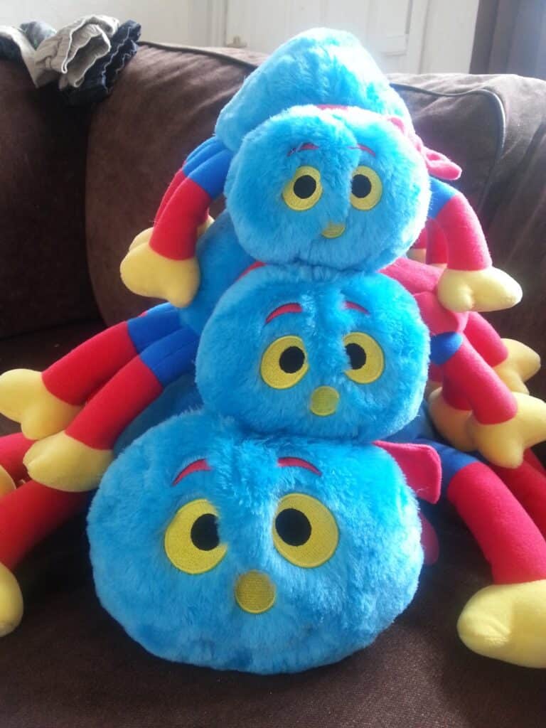 Three soft toys with round blue fluffy faces, big yellow and black eyes, red eyebrows, and long red, blue, and yellow legs are stacked on a brown sofa. There are clothes on the arm of the sofa and a door in the background.