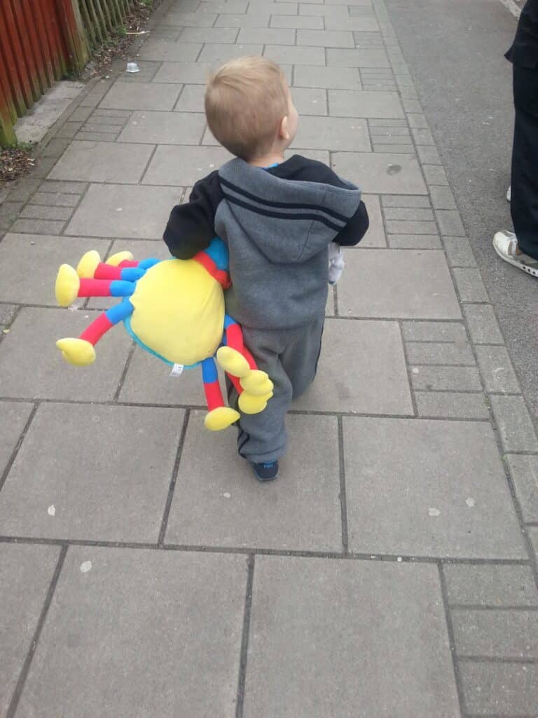 A young child in a grey tracksuit walks on a paved pavement, holding a large, colourful stuffed toy shaped like a spider with yellow, red, and blue limbs. The child is seen from behind, and part of an adult is visible ahead.