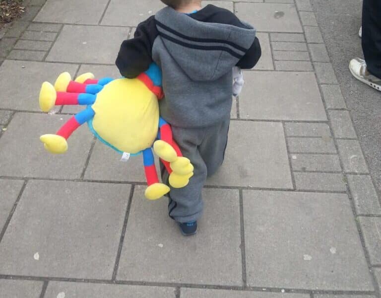 A small child in a grey tracksuit walks on a paved pavement, holding a colourful plush toy shaped like a spider or sun with blue, red, and yellow limbs. The view is from behind, showing the child’s back and part of another person ahead.