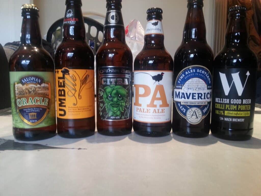 Six assorted beer bottles are lined up on a table. Each bottle has a unique label and branding, showing various names and designs, including Oracle, Umbel Ale, Citra, PA Pale Ale, Maverick, and a black and white labelled porter.