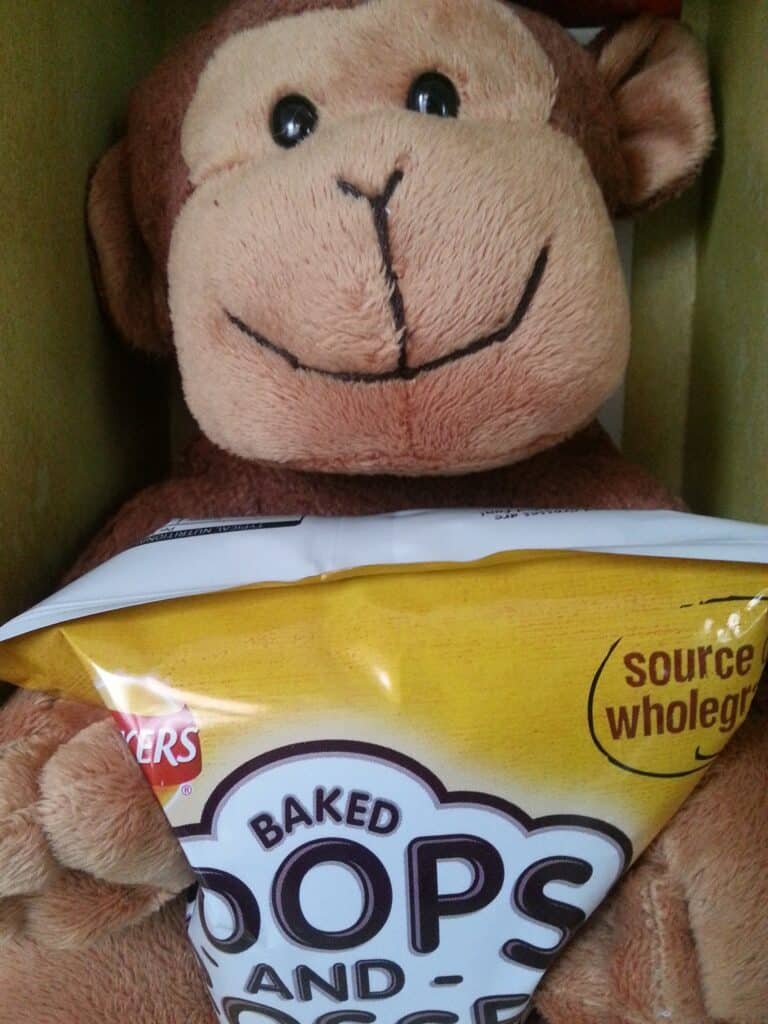 A plush brown monkey toy is sitting and holding a yellow bag of baked snack labelled "Baked Hoops and Crosses" with "source of wholegrains" printed on it. The monkey's face is cheerful, and the snack bag covers its lower body.