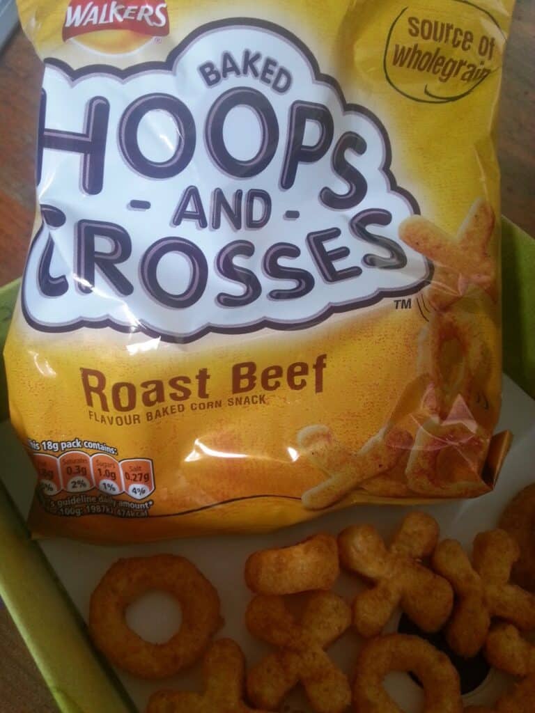 A yellow bag of Walkers Baked Hoops and Crosses Roast Beef flavour is shown, with some of the round “hoop” and “cross” shaped snacks scattered on a plate in front. The packet advertises that it is a source of wholegrain.
