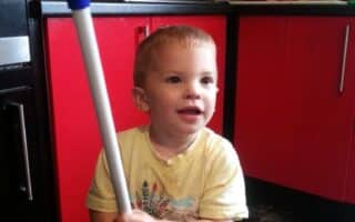 A young child with short fair hair, wearing a yellow t-shirt with a colourful print, stands in a kitchen holding a large duster with a white fluffy head and blue handle. The kitchen has red cupboards, black tile splashback, and various items on the worktops.