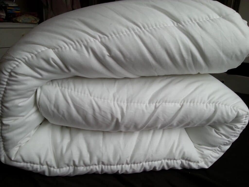 A thick, white, quilted duvet is folded neatly in thirds and placed on a dark surface. The fabric looks soft and plush, with visible stitching running across it in horizontal lines. The background is out of focus.