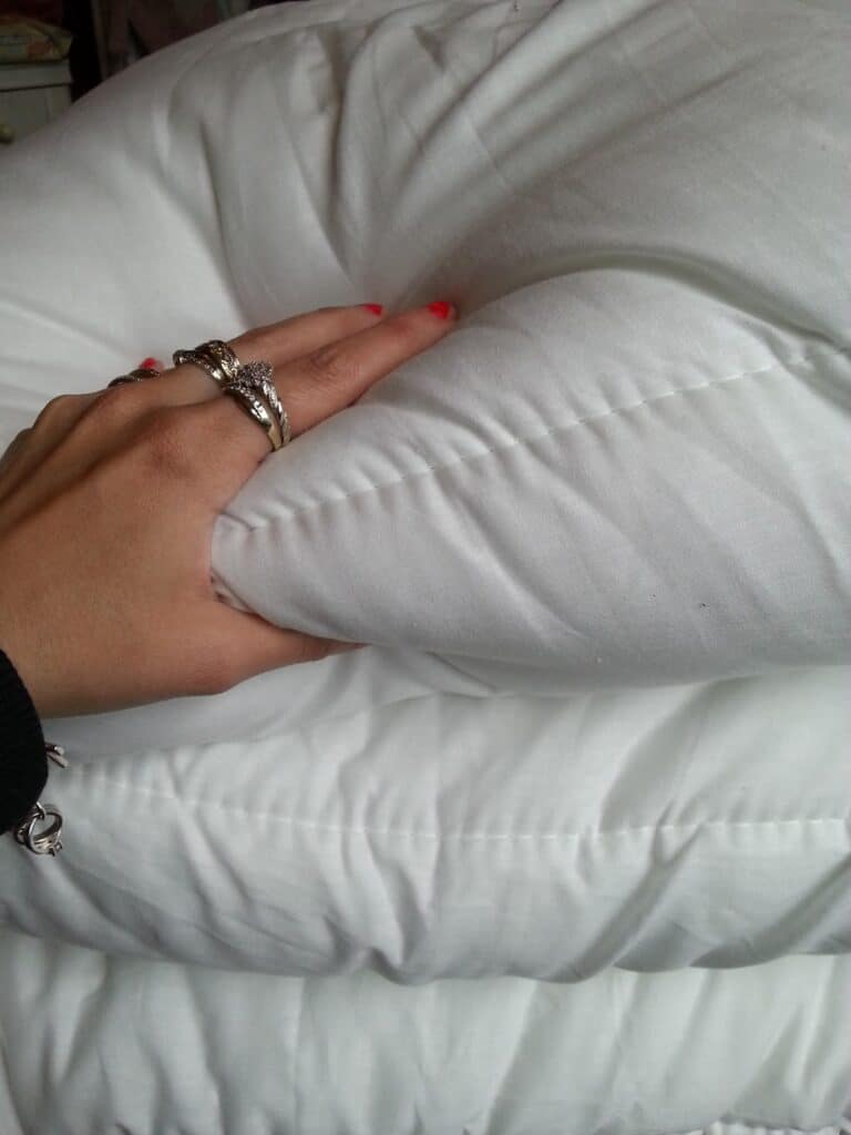 A hand with red-painted nails and wearing silver rings and a bracelet presses into a soft, white, quilted duvet or blanket, creating visible indentations and folds in the fabric. The bedding appears plush and clean.