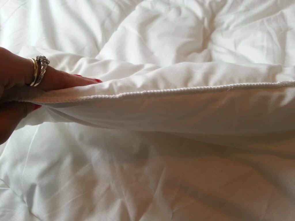A close-up of a hand holding up the edge of a white, quilted duvet or blanket, showing its thickness and soft texture. The hand has red-painted nails and is wearing silver rings. The background is a smooth, white bedding surface.