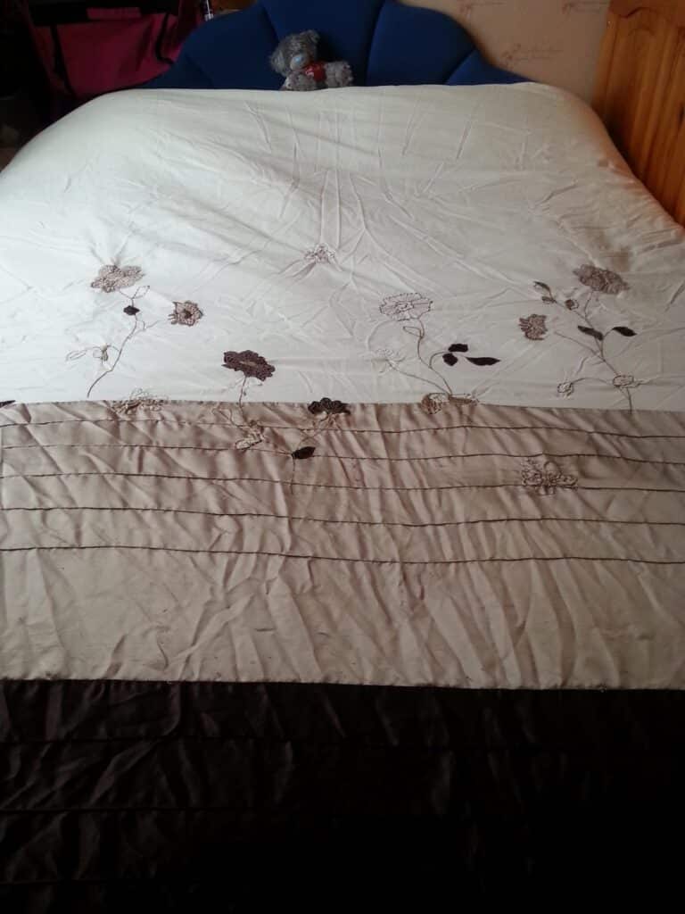 A bed covered with a white and beige quilt featuring floral embroidery. At the head of the bed is a blue cushion, and a small grey teddy bear sits propped up against it. The bedding has some creases and the room appears softly lit.