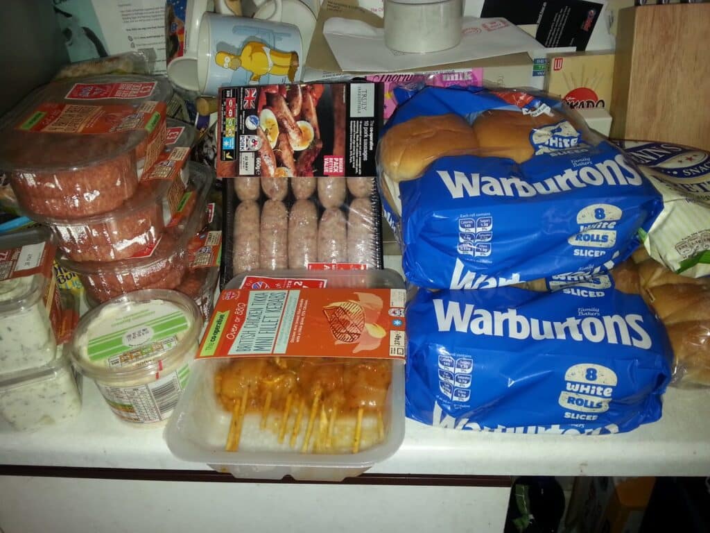 A kitchen worktop filled with food including packs of Warburtons white bread rolls, stacked beefburger patties, packs of sausages, tubs of dip or spread, skewered kebabs, sliced cheese, and various other groceries and items in the background.