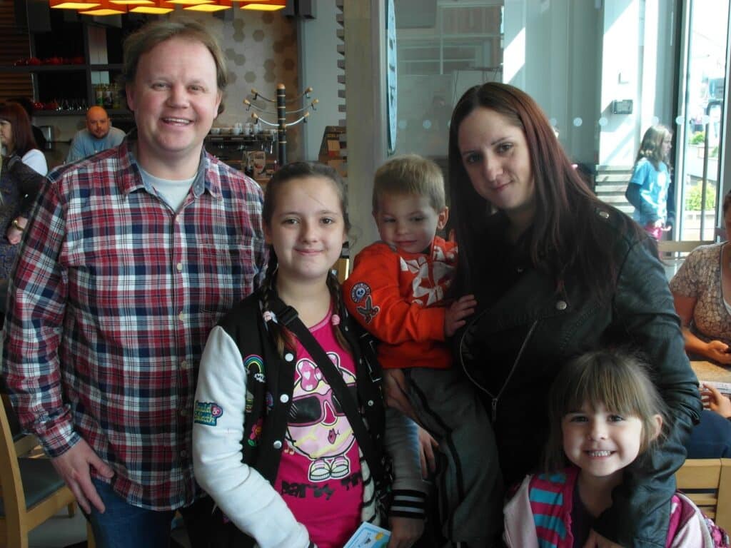 A smiling man in a checked shirt stands with a woman in a black jacket. They are with three children: two girls in colourful tops and a boy in an orange sweatshirt, indoors at a busy café with people and large windows in the background.