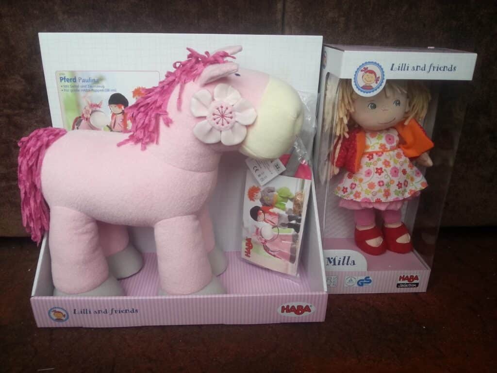 A pink plush horse with a flower on its ear sits in an open box next to a boxed soft doll with blonde yarn hair, wearing a red floral dress and orange cardigan. Both boxes are labelled "Lilli and friends" by HABA.