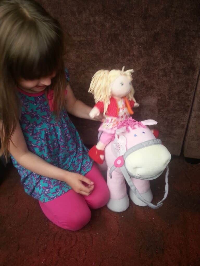 A young girl in a blue dress with pink leggings smiles and looks down as she places a blonde doll dressed in red and pink on top of a large pink plush hippopotamus toy. They are seated on a red carpet in front of a brown sofa.