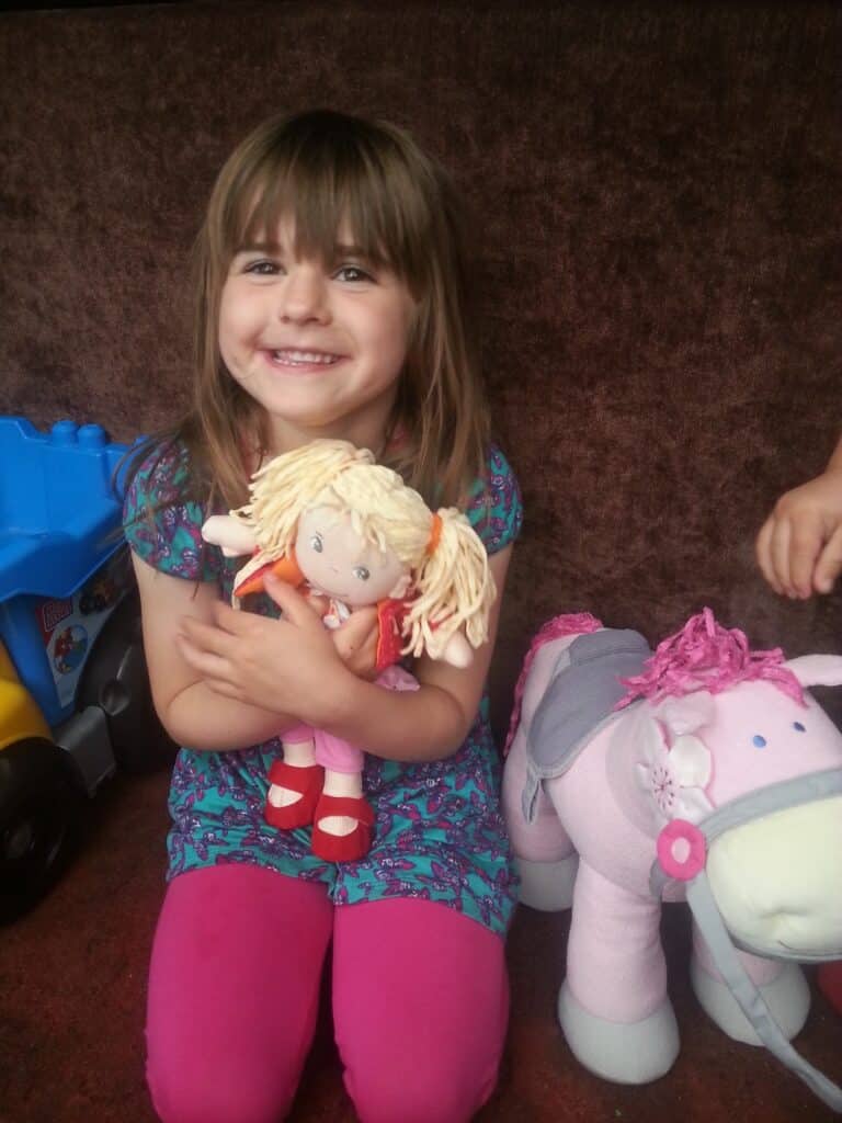 A young girl with brown hair and fringe, wearing a blue dress with a pink pattern and bright pink leggings, sits smiling and cuddling a blonde rag doll. A pink soft toy horse and a blue toy lorry are beside her against a brown background.