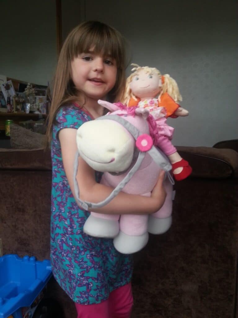 A young girl with long brown hair, wearing a blue dress with a purple pattern, smiles while holding a large plush pink horse. A blonde doll dressed in orange and pink sits on top of the horse. A brown sofa and some household items are in the background.