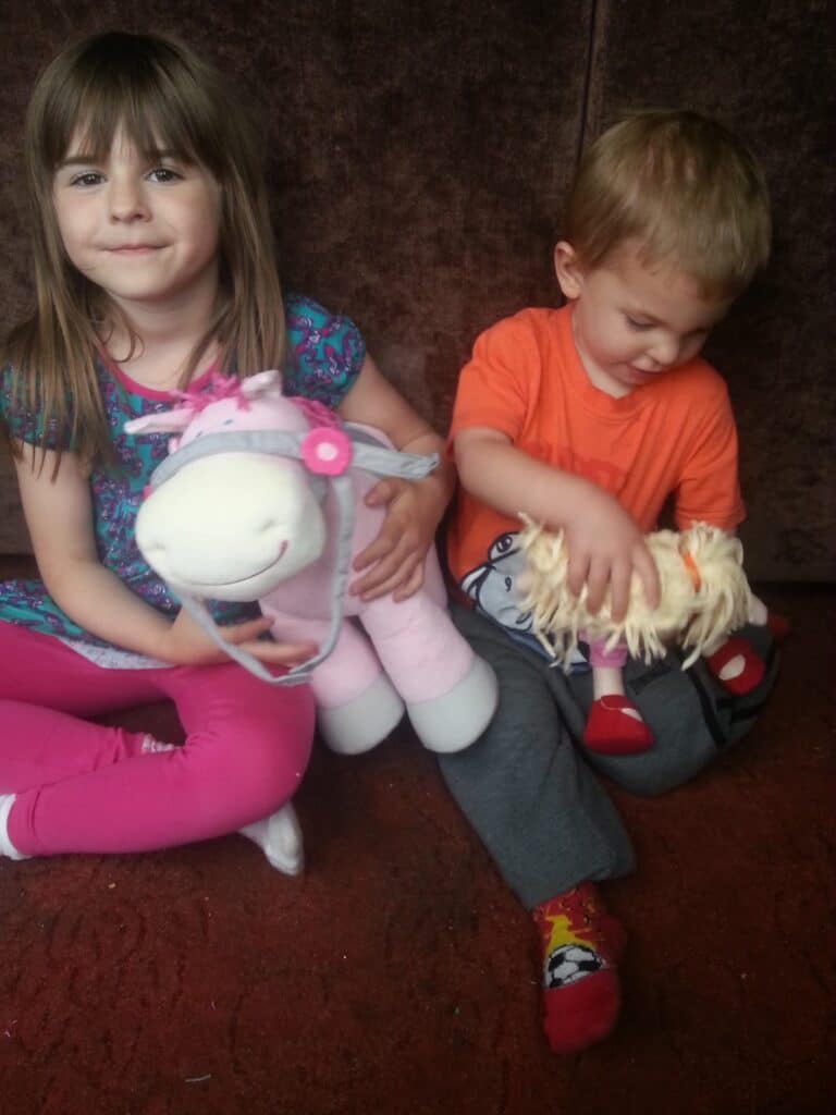 A young girl with brown hair in a blue floral top and pink leggings holds a large plush unicorn. Next to her, a young boy in an orange shirt and red socks looks down, holding a doll with woollen hair. They sit on a red carpet in front of a brown background.