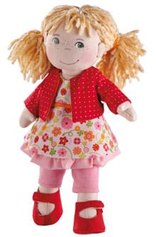 A soft fabric doll with yellow yarn hair in plaits, wearing a red polka-dot jacket, a floral dress with pink, yellow, and red flowers, pink leggings, and red shoes. The doll has stitched facial features and a friendly expression.
