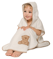 A young child sits wrapped in a cream-coloured hooded towel with beige trim. The towel features an embroidered brown teddy bear near the bottom. The child smiles and looks up, with the hood covering part of their head and bare toes visible.